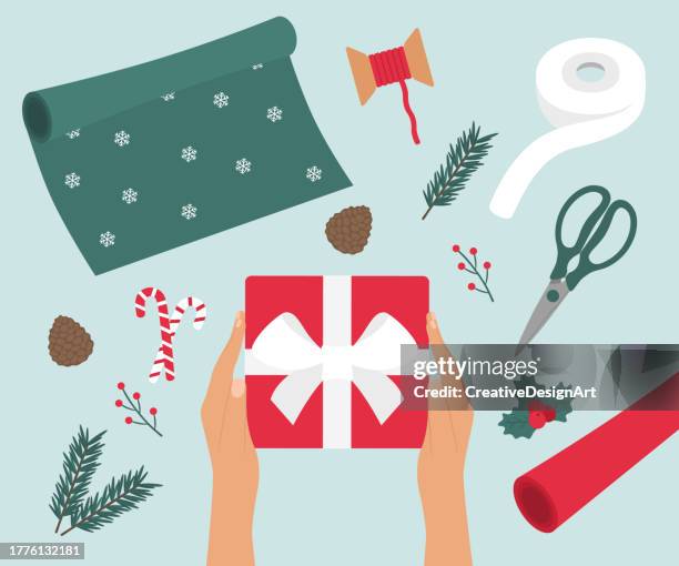 stockillustraties, clipart, cartoons en iconen met preparing christmas present. high angle view of table with fir tree branches, pine cones, wrapping papers, scissors and ribbon spool. hands holding gift box. christmas celebration concept - kado inpakken