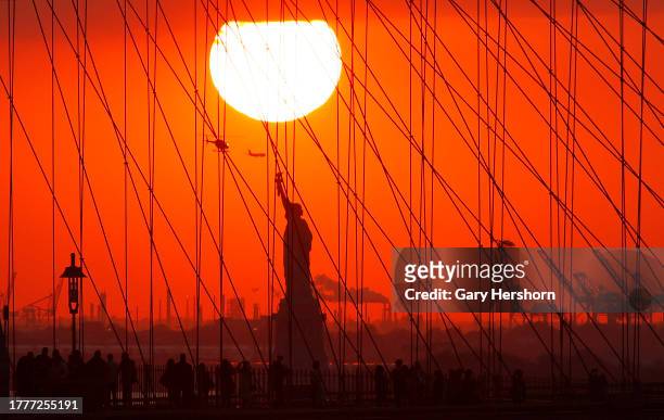People walk across the Brooklyn Bridge as the sun sets behind the Statue of Liberty on November 5 in New York City.