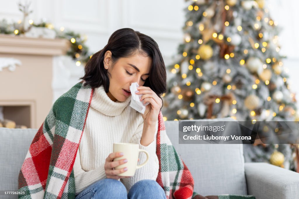 A young woman fell ill during the Christmas and New Year holidays. She sits at home on the sofa covered with a blanket, wipes her nose from a runny nose with a napkin, holds a cup of drink, medicine