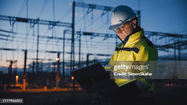 ingeniero eléctrico masculino maduro dedicado usando computadora portátil en la planta de energía oscura durante el crepúsculo - producción de combustible y energía fotografías e imágenes de stock
