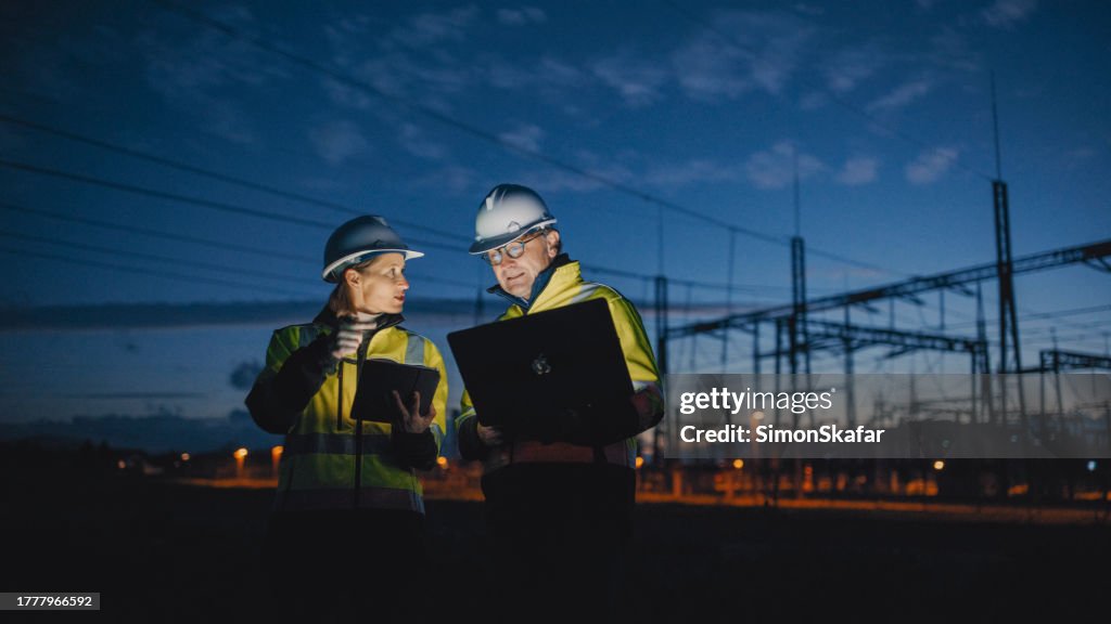Elektroingenieure diskutieren über Laptop und digitales Tablet, während sie nachts im dunklen Kraftwerk stehen