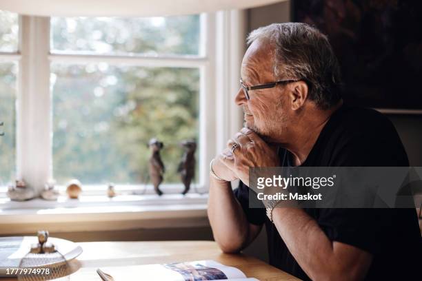 senior man with hand on chin looking through window at home - tisch betrachten stock-fotos und bilder