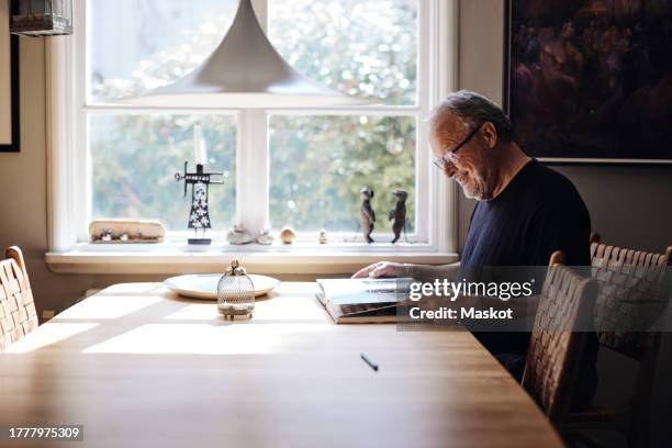 smiling senior man with eyeglasses looking at picture book on table at home - accessory stockfoto's en -beelden