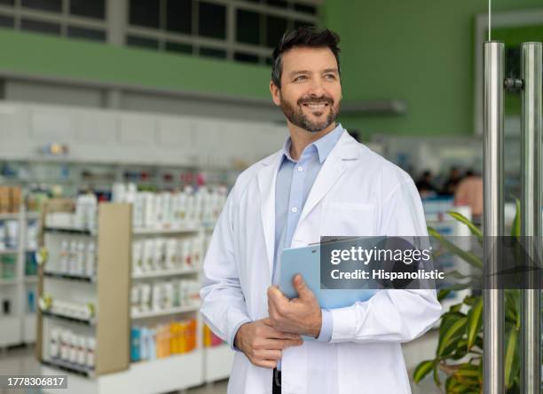 happy pharmacist standing at the door of the drugstore holding a clipboard - looking away stock pictures, royalty-free photos & images