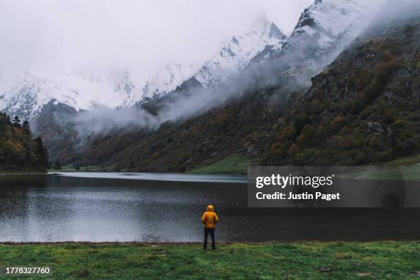 dramatic view of an unrecognisable person in a yellow puffer coat looking over the beautiful lac d'estaing in the french pyrenees mountains - puffer jacket stock pictures, royalty-free photos & images