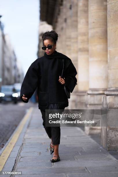 Ellie Delphine wears Celine sunglasses, a black oversized wool scarf, a black coat from Toteme, black cropped pants, a heart shaped bag from Alaia,...
