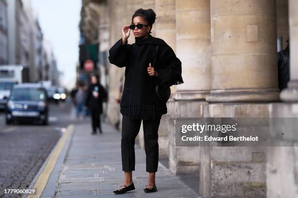 Ellie Delphine wears Celine sunglasses, a black oversized wool scarf, a black coat from Toteme, black cropped pants, a heart shaped bag from Alaia,...