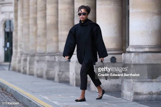 Ellie Delphine wears Celine sunglasses, a black oversized wool scarf, a black coat from Toteme, black cropped pants, a heart shaped bag from Alaia,...