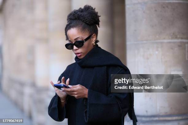 Ellie Delphine wears Celine sunglasses, a black oversized wool scarf, a black coat from Toteme, black cropped pants, during a street style fashion...