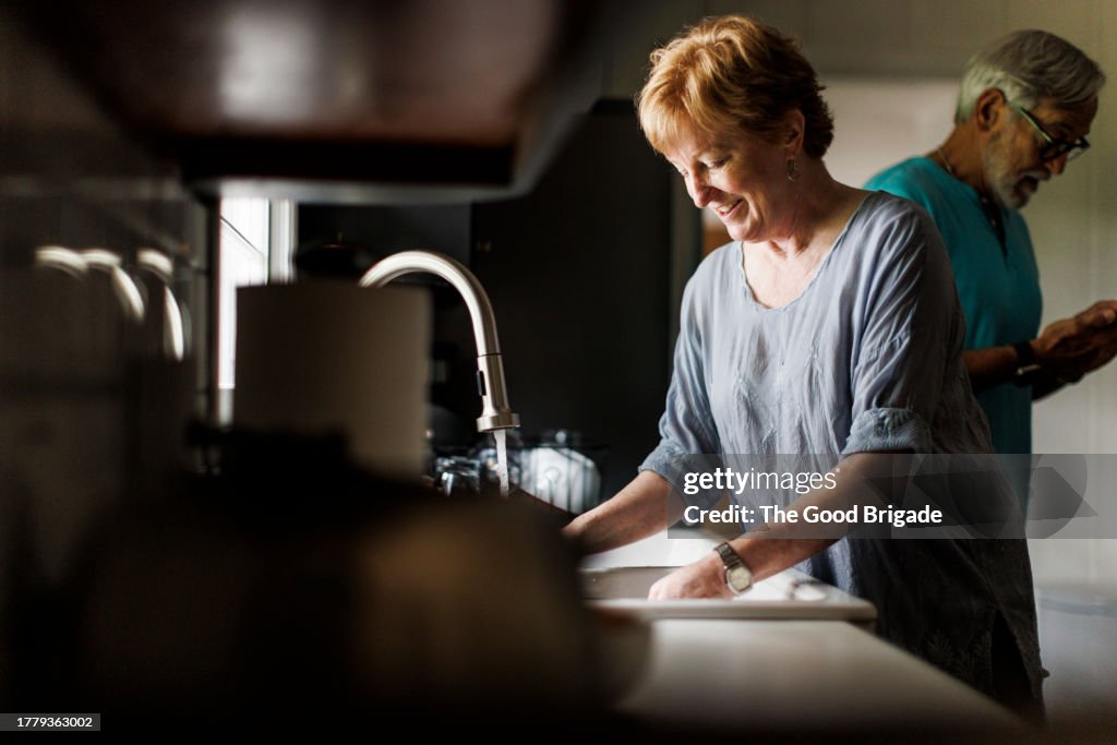 Smiling senior woman using sink while man standing behind kitchen