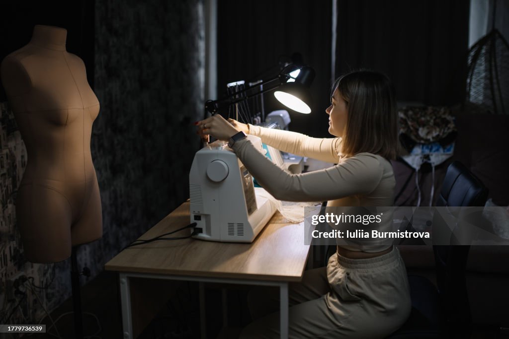 Skilled female tailor sits at dimly lit table navigating contours of sewing machine beneath focused beam of electric lamp