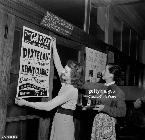 Barmaid Pat Barclay puts up a poster advertising The Castle pub's jazz night on Tooting Broadway, London, April 1954. At right is a woman named as...