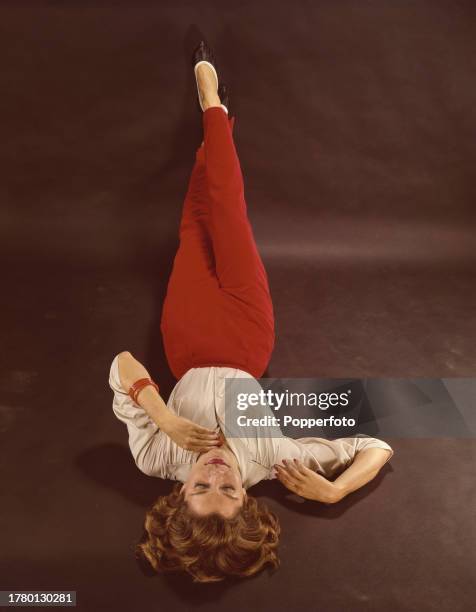 Posed studio portrait of a female fashion model wearing a cream blouse and bright red slacks, she lies on the floor of the studio with raised legs,...