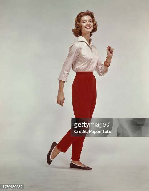 Posed studio portrait of a female fashion model wearing a cream blouse, bright red slacks and black ballet pumps, London, 14th October 1961.