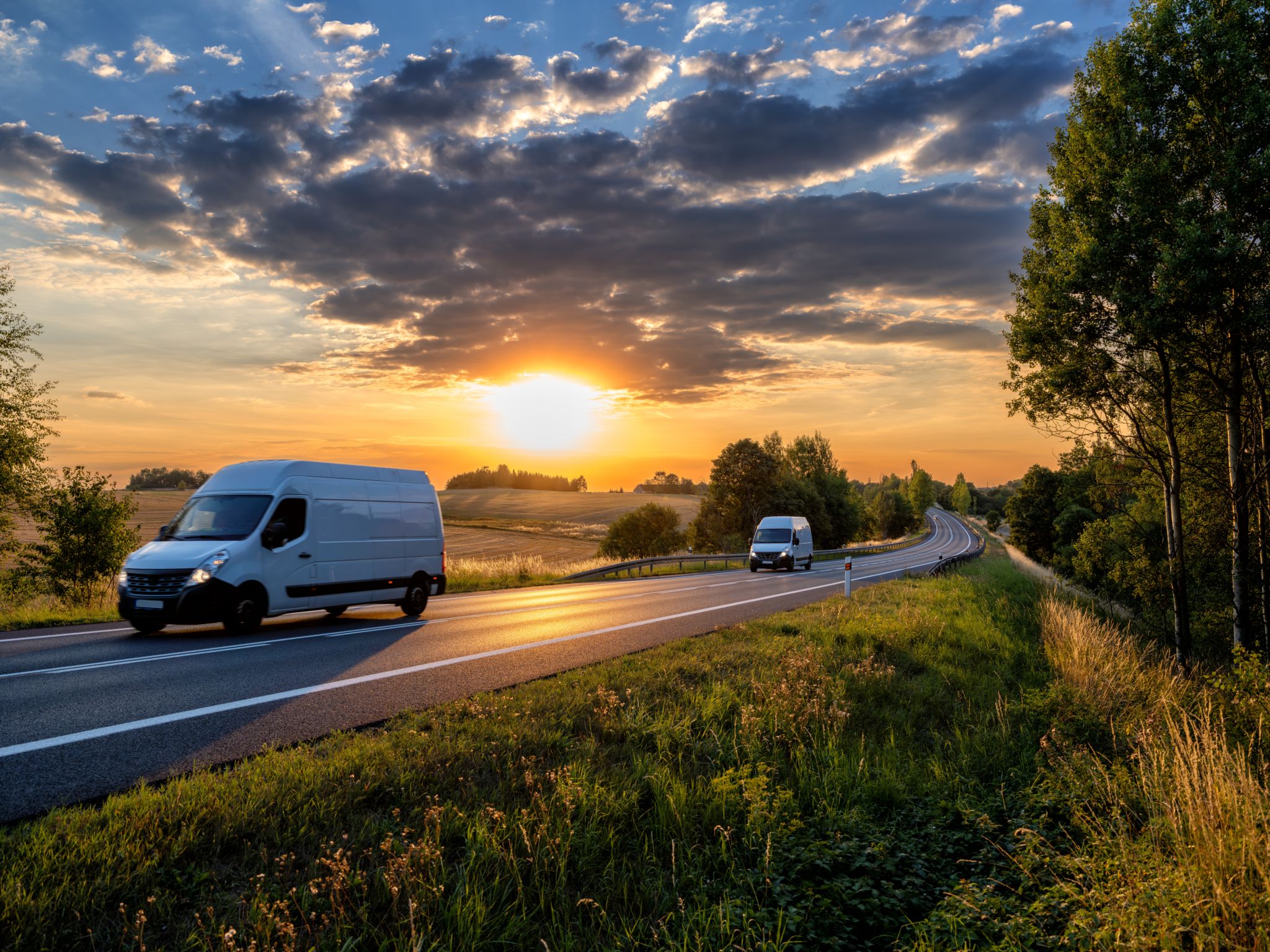 Two white fast delivery vans driving on the asphalt road in rural landscape at sunset with dark cloud Two white fast delivery vans driving on the asphalt road in rural landscape at sunset with dark cloud