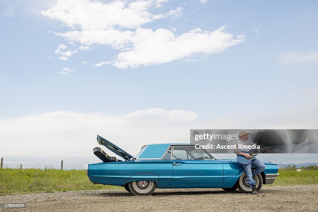 Man rests against older model car, plays guitar