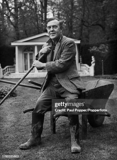 British fashion designer Norman Hartnell, wearing a tweed jacket and gumboots as he sits on the edge of a wheelbarrow, in the garden of his home,...