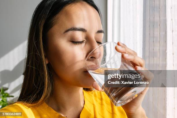 young latin american woman drinking a glass of fresh water standing close to the window in the morning. healthy lifestyle and people concept. - trinken stock-fotos und bilder