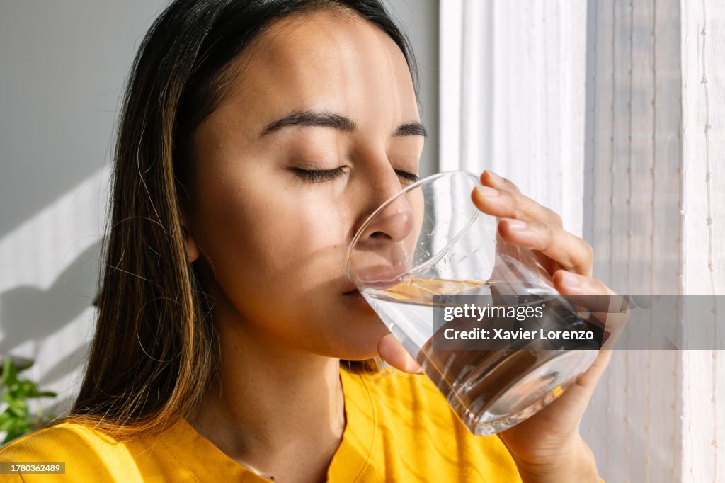 Young Latin American woman drinking a glass of fresh water standing close to the window in the morning. Healthy lifestyle and people concept.