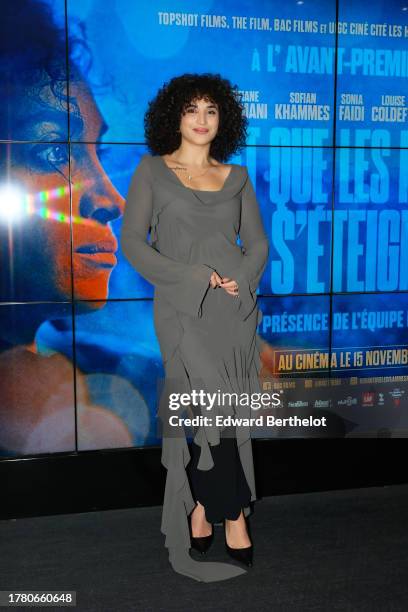 Camelia Jordana wears a necklace, a gray low neck flowing dress with gathered low parts and flared oversized sleeves, black pointed shoes, attends...