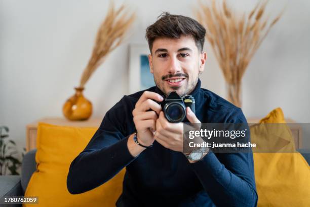 portrait of a handsome young man holding a camera while sitting on a sofa at home - facing things head on stock pictures, royalty-free photos & images