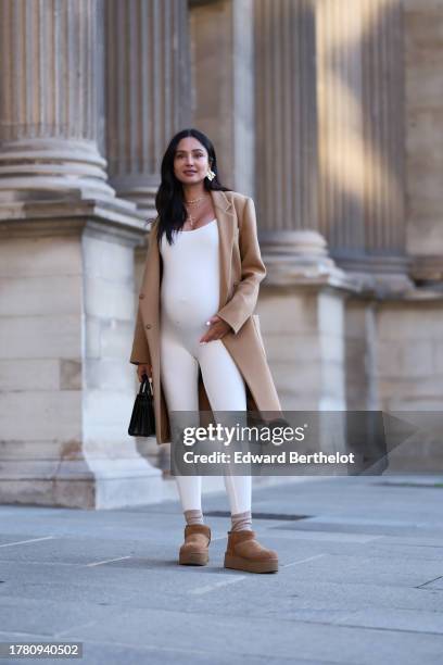 Patricia Gloria Contreras wears golden earrings, a necklace, a pale brown / beige winter coat by Saint Laurent YSL, a white low neck jumpsuit by...