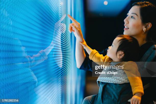 young asian mother and baby girl touching illuminated led digital display screen. mother and her kid's finger pointing at blue coloured digital display in the dark. connecting to the future. futuristic concept. innovation, lifestyle and technology concept - orient photos et images de collection