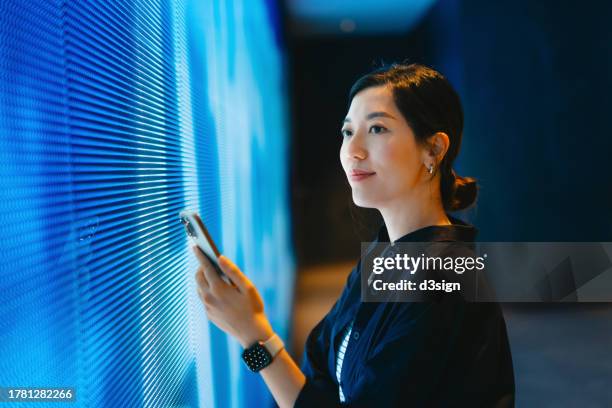 confident young asian woman using smartphone against blue coloured illuminated led digital display screen in the dark. connecting to the future. futuristic. artificial intelligence. innovation, lifestyle and technology. smart banking with technology - pantalla de proyección fotografías e imágenes de stock