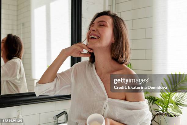 woman laughing while applying cream while standing in the bathroom - head and shoulders stock-fotos und bilder