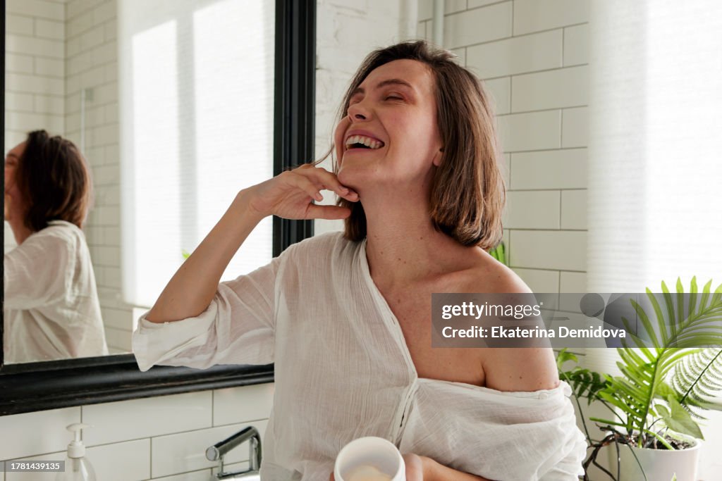 Woman laughing while applying cream while standing in the bathroom