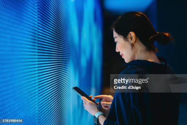 confident young asian woman using smartphone against blue coloured illuminated led digital display screen in the dark. connecting to the future. futuristic. artificial intelligence. innovation, lifestyle and technology concept - technology security stock pictures, royalty-free photos & images