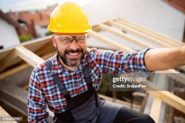 mid-adult caucasian male roofer taking a break from work, while sitting on a roof beam and taking selfie - dakdekker stockfoto's en -beelden