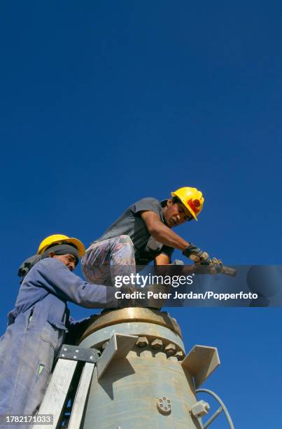 Two workers, wearing Bechtel Corporation hard hats, tighten bolts on pipework during construction of the Maghreb-Europe Gas Pipeline in the desert...