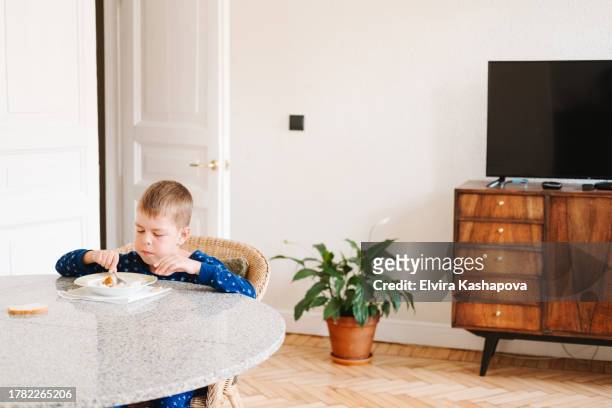 9-year-old boy has a breakfast of rice porridge and nuggets in the dining room with a hearing aid in his ear. schoolchild having breakfast at home against the background of a tv and a flower in a pot - eating nuggets ストックフォトと画像