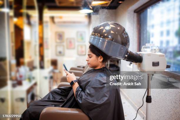 woman using mobile phone while getting hair treatment under a professional hair steamer - haarbehandeling stockfoto's en -beelden