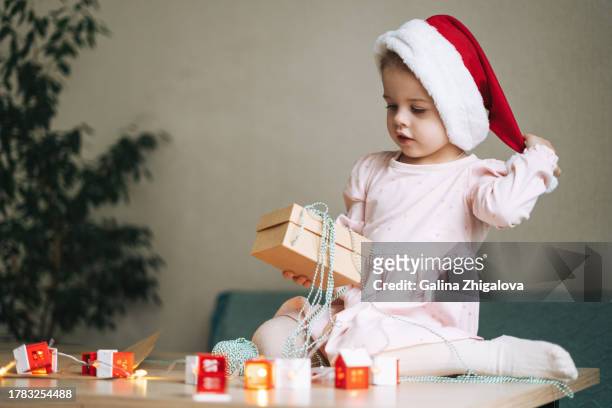 cute little girl in santa hat opening gift box at the table at home in christmas time - baby 3 months stock-fotos und bilder