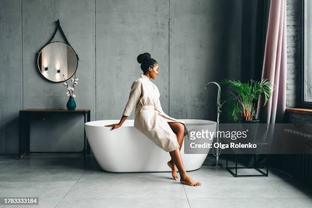 spa day. relaxed woman in robe sit on bathtub and look to the window - bath robe stockfoto's en -beelden