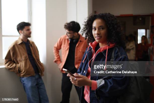 a female student posing with her phone in hands and smiling in the corridors of college - métis stock pictures, royalty-free photos & images