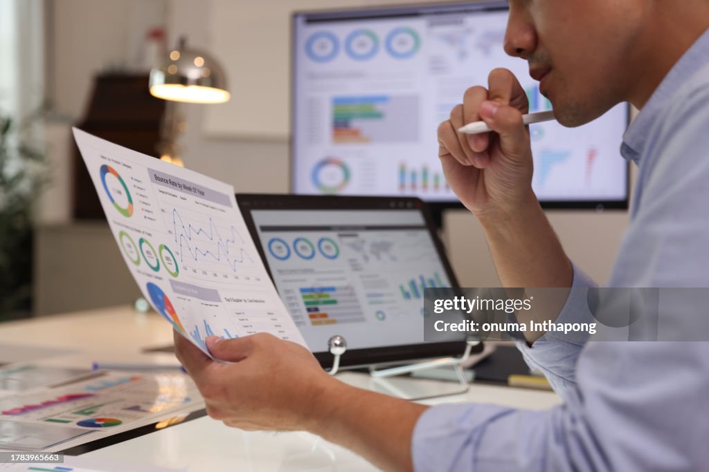 Focused businessman analyzing marketing reports on a desktop monitor and reviewing paper graphs, financial stats, and startup project infographics.