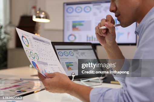 Focused businessman analyzing marketing reports on a desktop monitor and reviewing paper graphs, financial stats, and startup project infographics. Focused businessman analyzing marketing reports on a desktop monitor and reviewing paper graphs, financial stats, and startup project infographics.