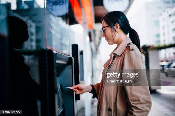 young asian woman inserting her bank card into automatic cash machine on city street. withdrawing money. paying bills. checking account balances. making a bank transfer. privacy protection. smart banking on the go - atm stock pictures, royalty-free photos & images