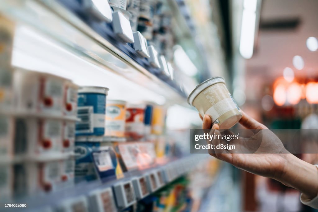 Close-up of a woman's hand shopping for fresh organic healthy yoghurt along the dairy aisle in supermarket, reading the nutrition label on the pot. Routine grocery shopping. Healthy eating lifestyle. Making healthier food choices. Smart eating concept