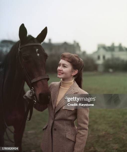 British actress Janette Scott, wearing a tweed riding jacket over a yellow turtleneck sweater, as she stands smiling beside a horse which she holds...