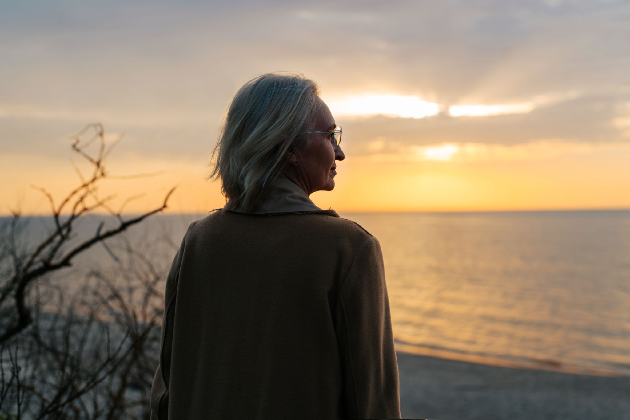 Senior caucasian woman wearing glasses and a coat standing on the seashore at sunset and looking away. Rear view of a pensive age lonely lady at the sea in the evening Senior caucasian woman wearing glasses and a coat standing on the seashore at sunset and looking away. Rear view of a pensive age lonely lady at the sea in the evening