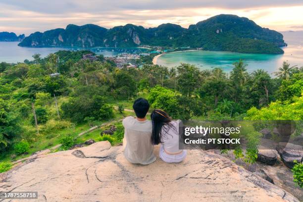 a man and woman sit on a viewpoint on phi phi island overlooking the city, mountains and sea at phi phi island, krabi, thailand. - provincia-di-phuket foto e immagini stock