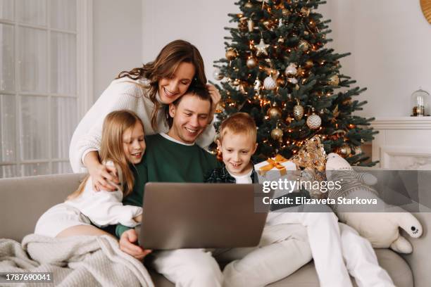 a happy family with two teens children in casual clothes hugging, having fun and chatting in the living room with a sofa and a decorated christmas tree in the bright interior of the house during the christmas holidays. - happy holidays family stock pictures, royalty-free photos & images