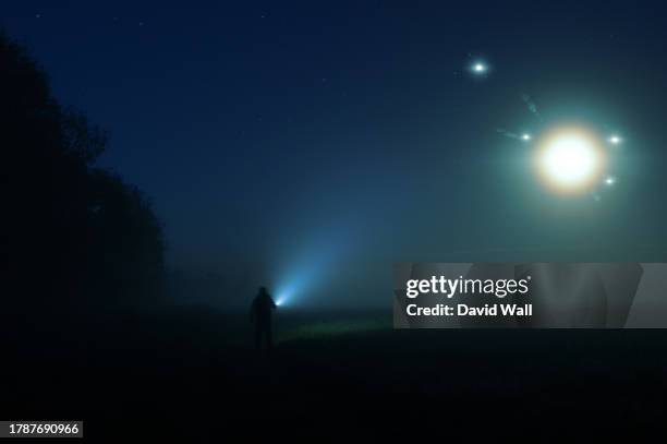 a man with a torch standing in the countryside on a misty summer night. with glowing ufo lights floating in the sky. - ovni fotografías e imágenes de stock