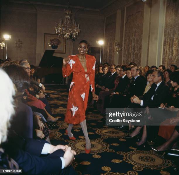 Fashion model wearing a red dress with white butterfly motifs, waves as she walks between two rows of seated guests at a fashion show, circa 1975.