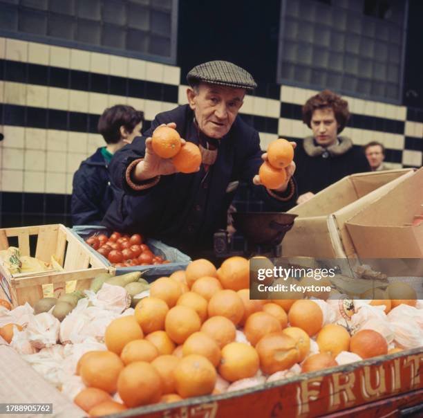 Market trader poses with four oranges in his hands as he stands at his greengrocer stall, laden with crates of fresh produce, with shoppers in the...