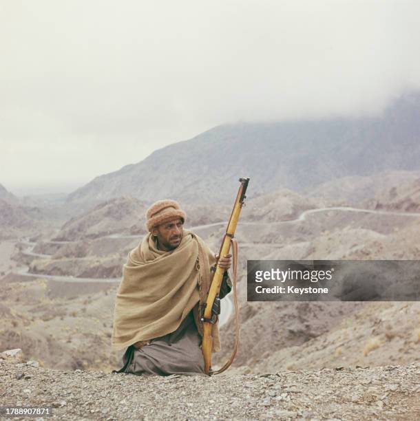 Man wrapped in a shawl and wearing a pakol, holding an Ishapore Number 4 Mk1 rifle as he sits in the rugged terrain of the Khyber Pass, a mountain...